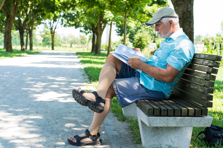 Side view portrait of senior man with cap on his head, short pants and compression socks for varicose veins in park, reading a book while sitting in shadow on hat summer day.