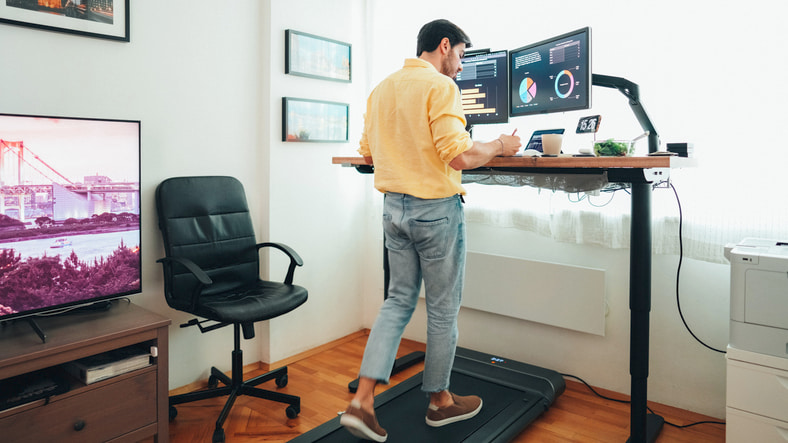 Man working from home, using a standing desk with a treadmill underneath to stay active while working.