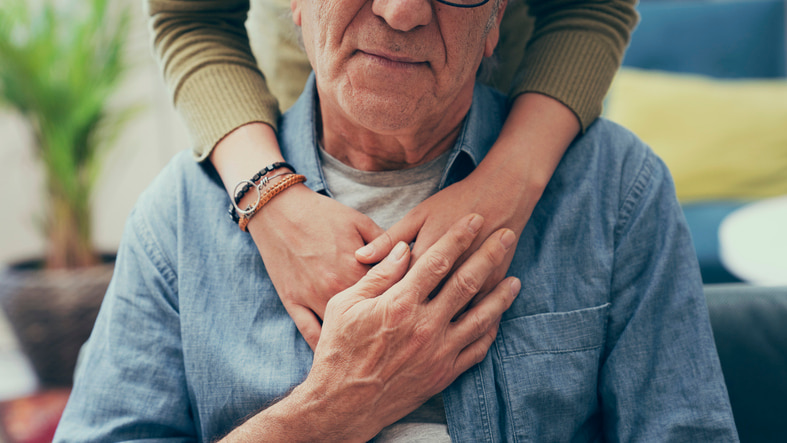 Senior man with hand on heart and person hugging him from behind