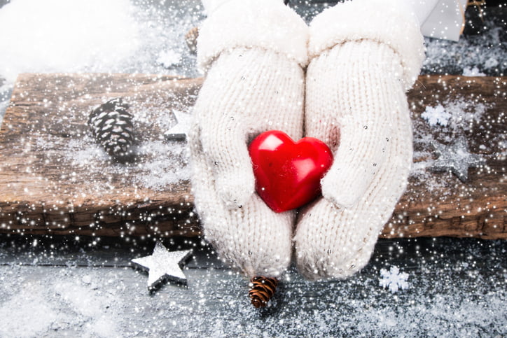 Two hands in white mittens holding a red heart in a snowy background