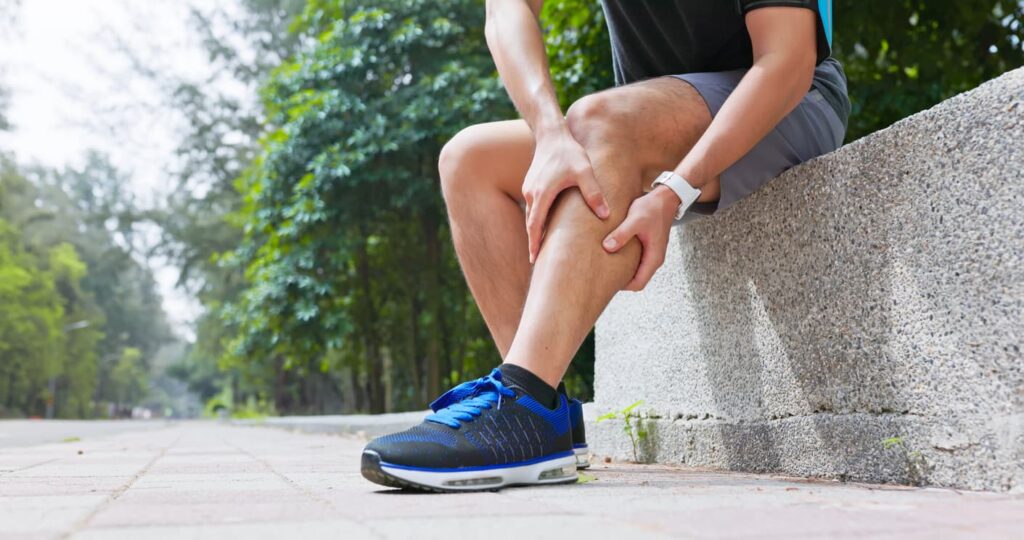 exercise hurt - closeup young man is sporting in park he stops and sits down by roadside chair suffers from calf cramping with hands touching massage the painful point straightening legs pull foot for ease