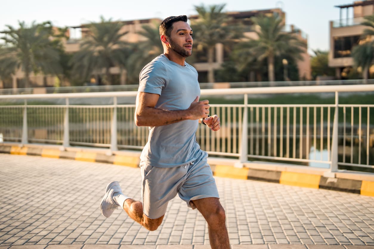 A mid-adult man in athletic attire runs outdoors on a paved pathway at sunrise, surrounded by palm trees and modern buildings.